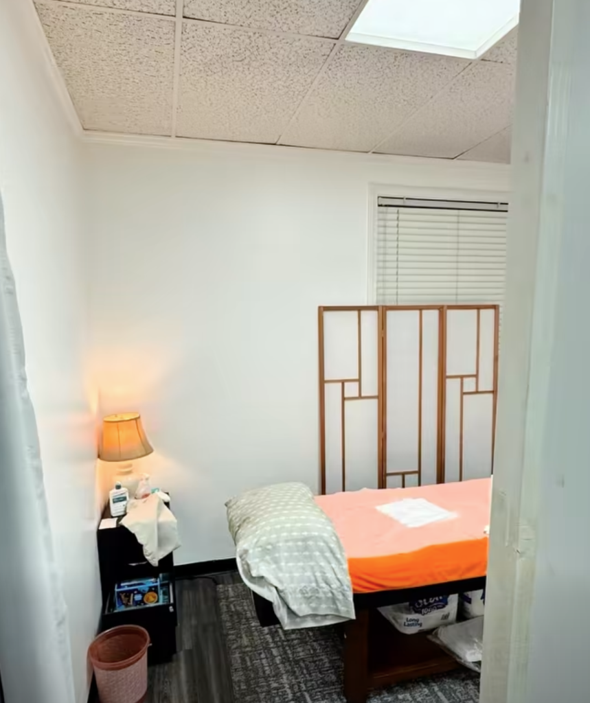 Massage room with a wooden partition, a massage table with orange linens, a side table with a lamp, and a window with blinds.
