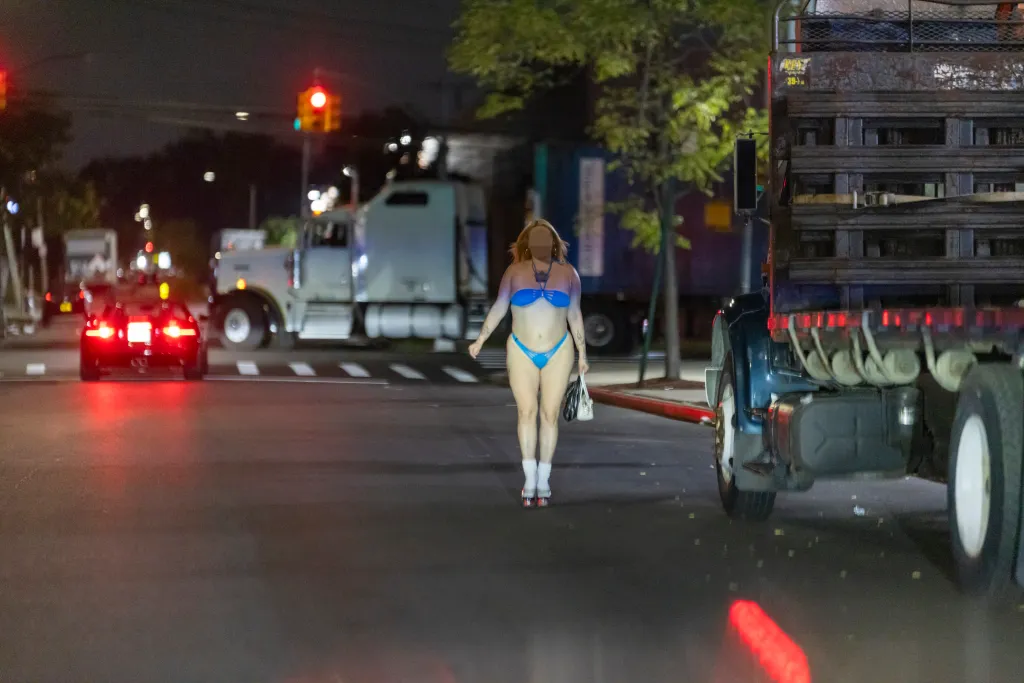 A woman wearing a blue bikini and white socks walking on a street at night near a large truck and cars in East New York.
