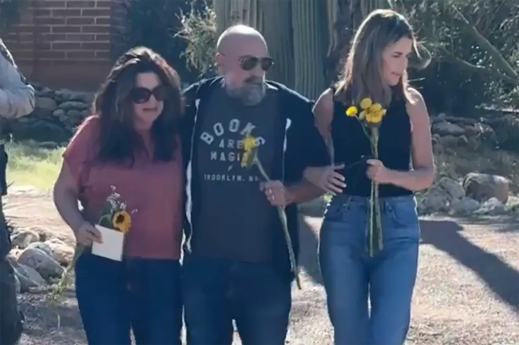 A man holding a single yellow flower walks between two women, each holding flowers, on a sunny path.