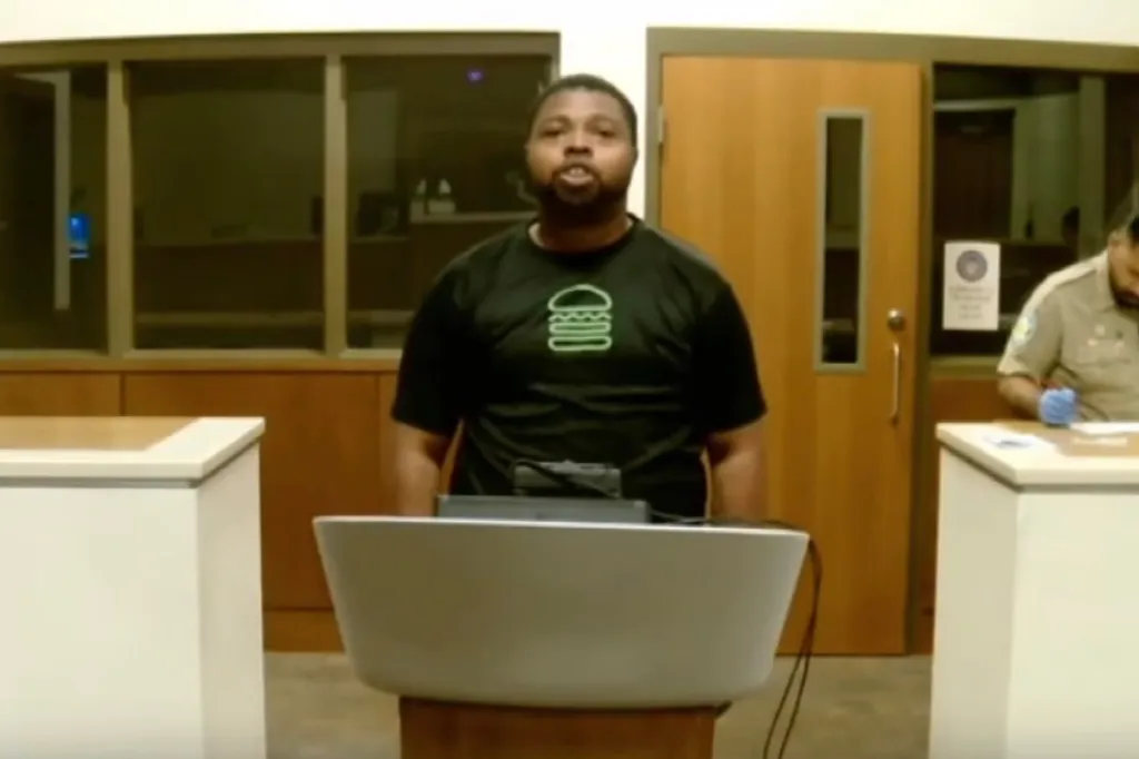 A man in a black Shake Shack t-shirt speaks from a podium with an officer seated to his right.