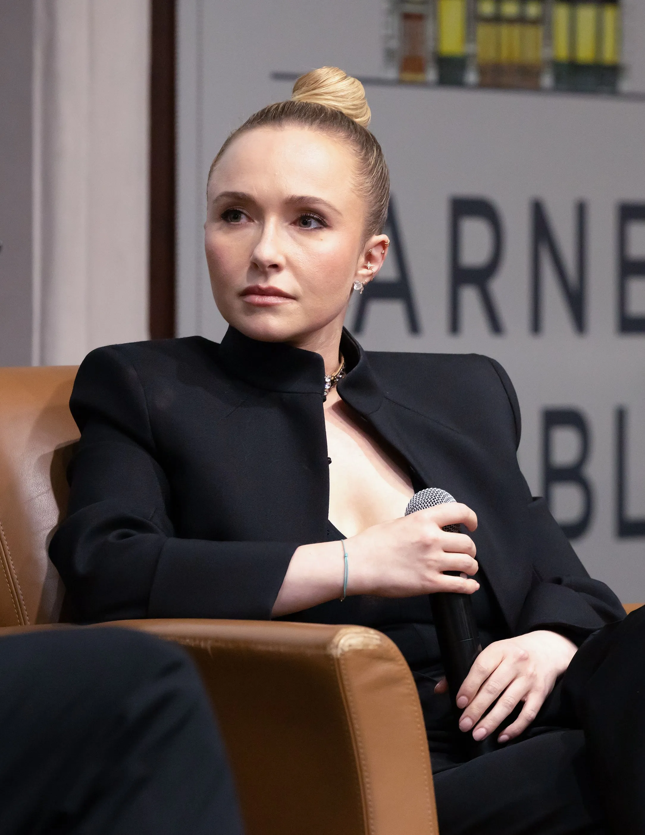 Hayden Panettiere in a black pantsuit, seated on a brown leather chair, holding a microphone.