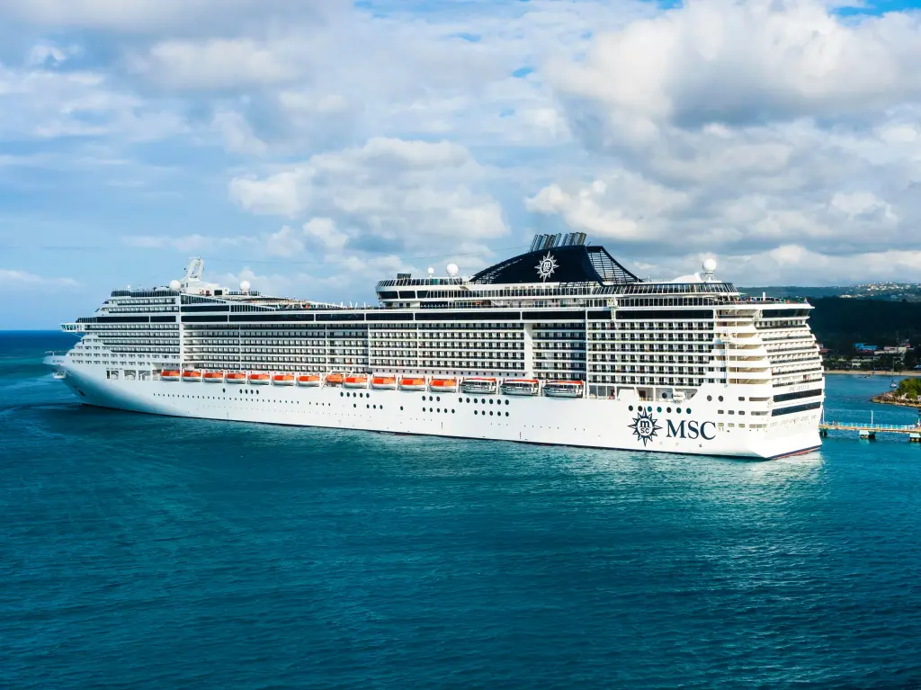 A large white MSC cruise ship with lifeboats on its side docked next to a pier, with a coastline visible in the background under a cloudy sky.