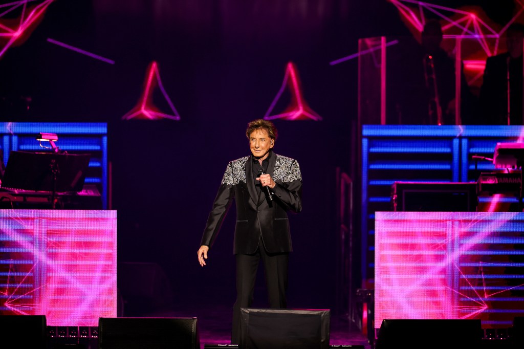 Barry Manilow performing onstage with a microphone in a black suit with embroidered shoulders, against a dark background with pink and blue geometric lights.