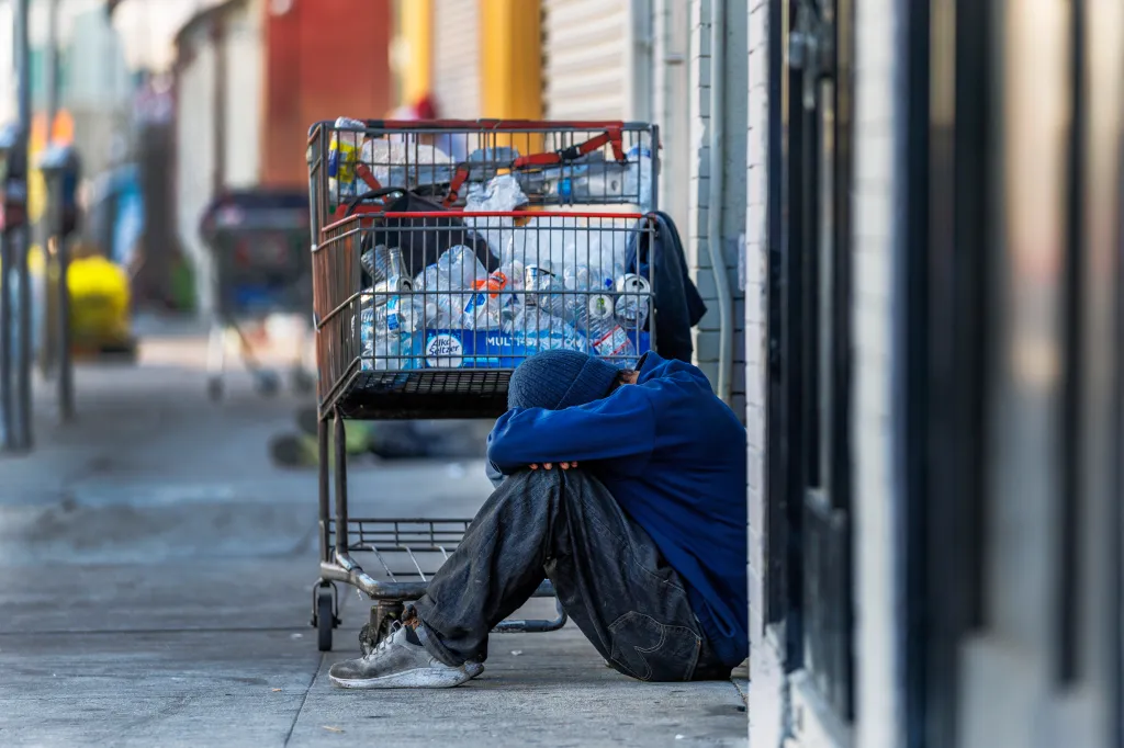 A homeless man leans his head on his knees next to a grocery cart filled with empty water bottles.