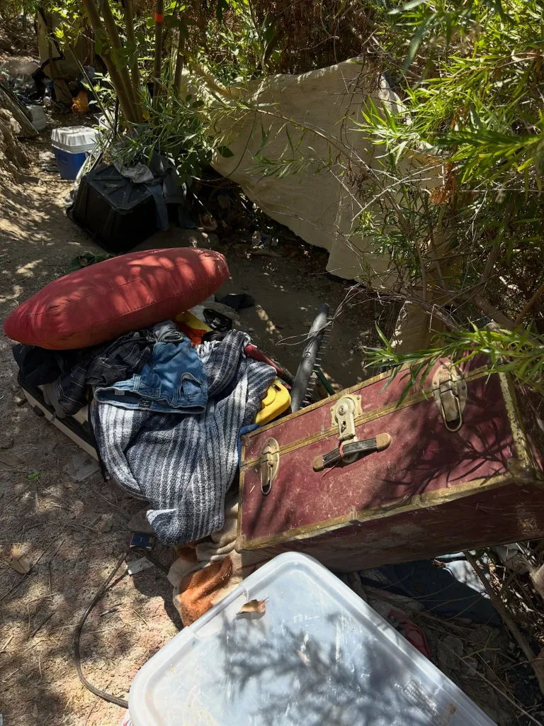 Personal belongings and debris lay on the ground in a homeless encampment.