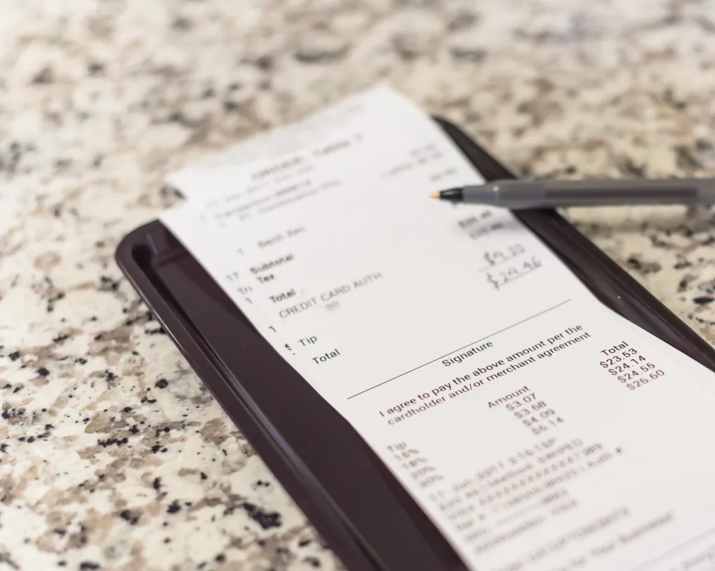 Restaurant bill with handwritten total, suggested tips, and a pen on a marble table.