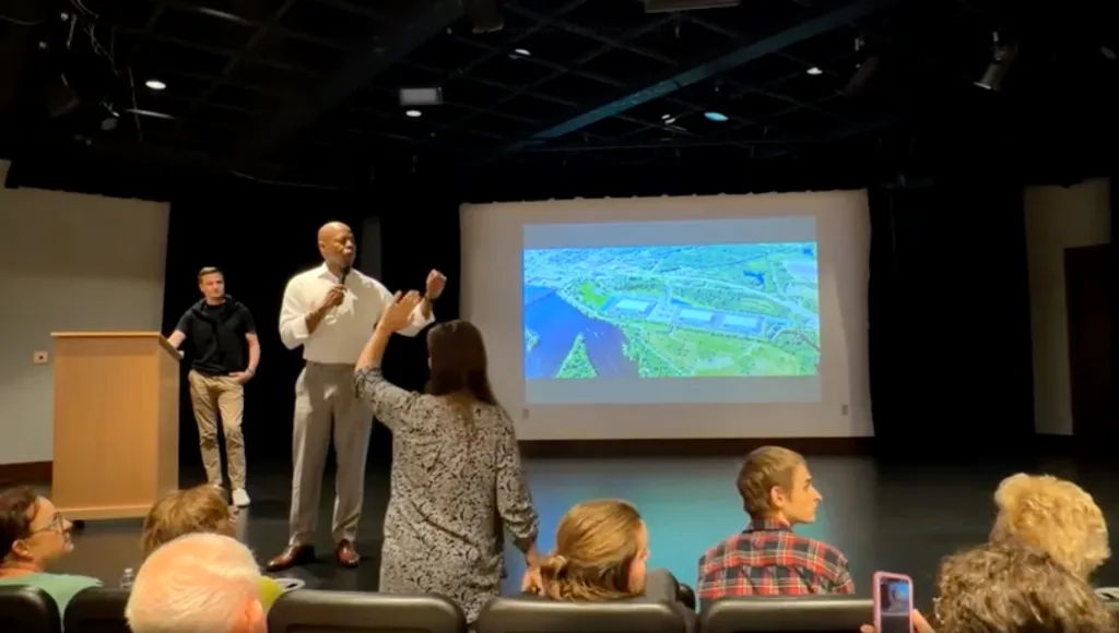 Eric Adams presents at a data center pitch with an aerial view of the proposed site on screen, while being heckled by an audience member.