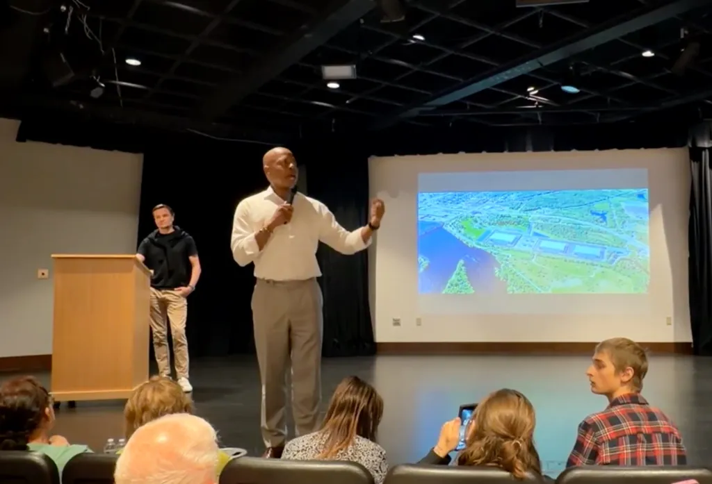 Eric Adams speaking at a data center pitch with an aerial view of a data center projected behind him.