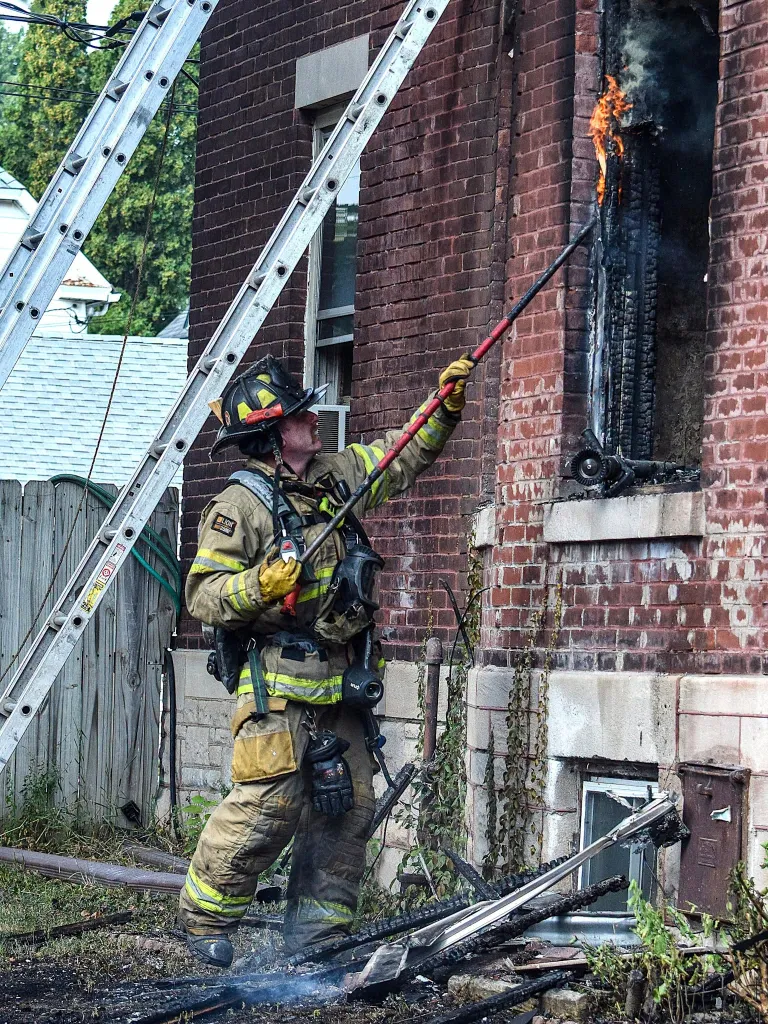 A firefighter uses a pole to suppress flames coming from a window of a brick building.