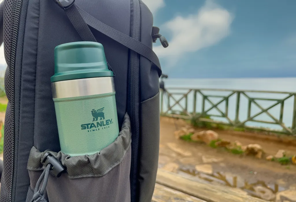 A green Stanley Action Trigger thermos in the side pocket of a black backpack, with an ocean view in the background.