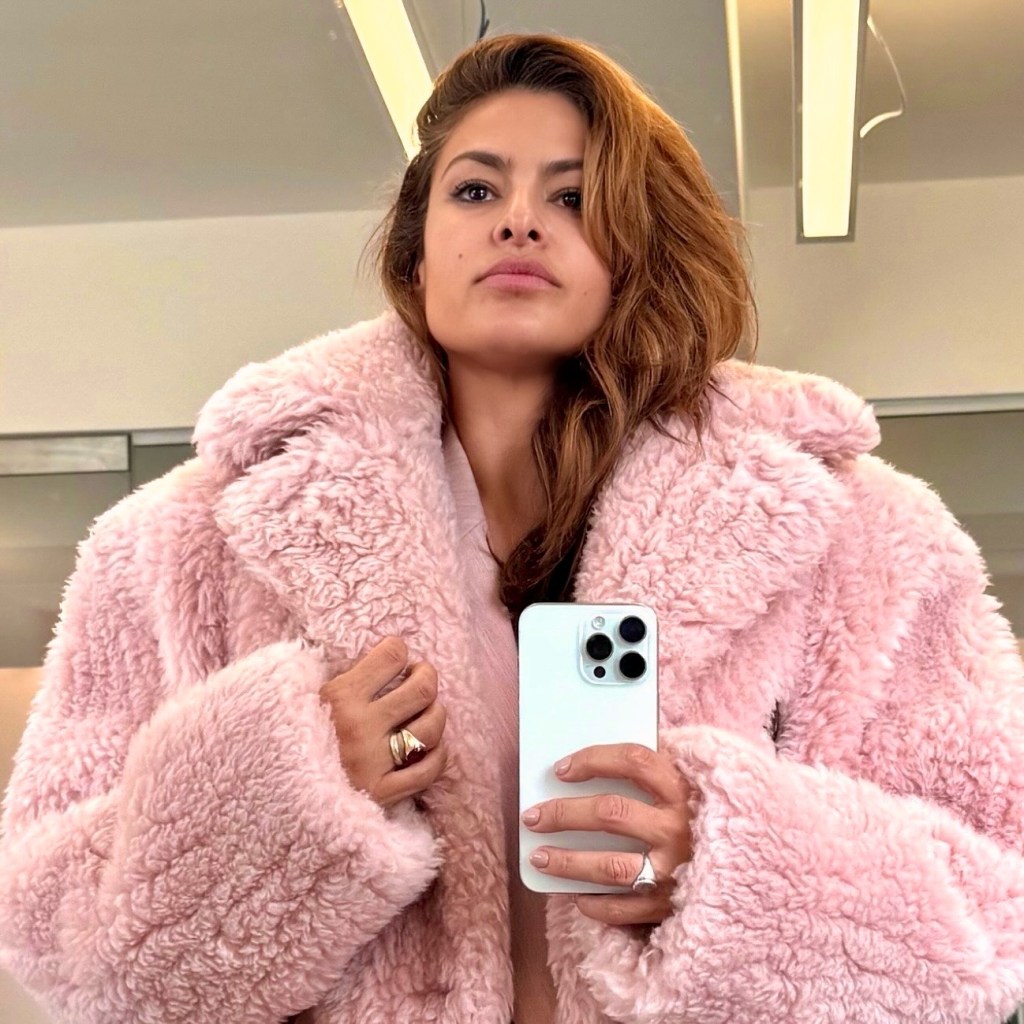 Eva Mendes wearing a pink fluffy coat, holding a white smartphone to take a selfie.
