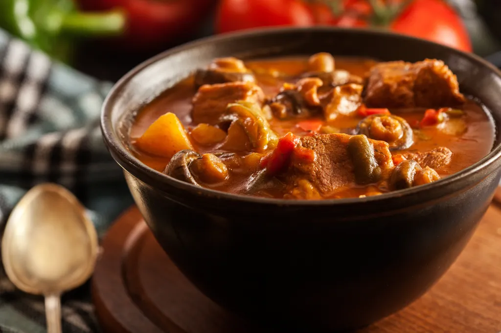 Stew soup with meat, potatoes, mushrooms, and red pepper in a dark bowl.