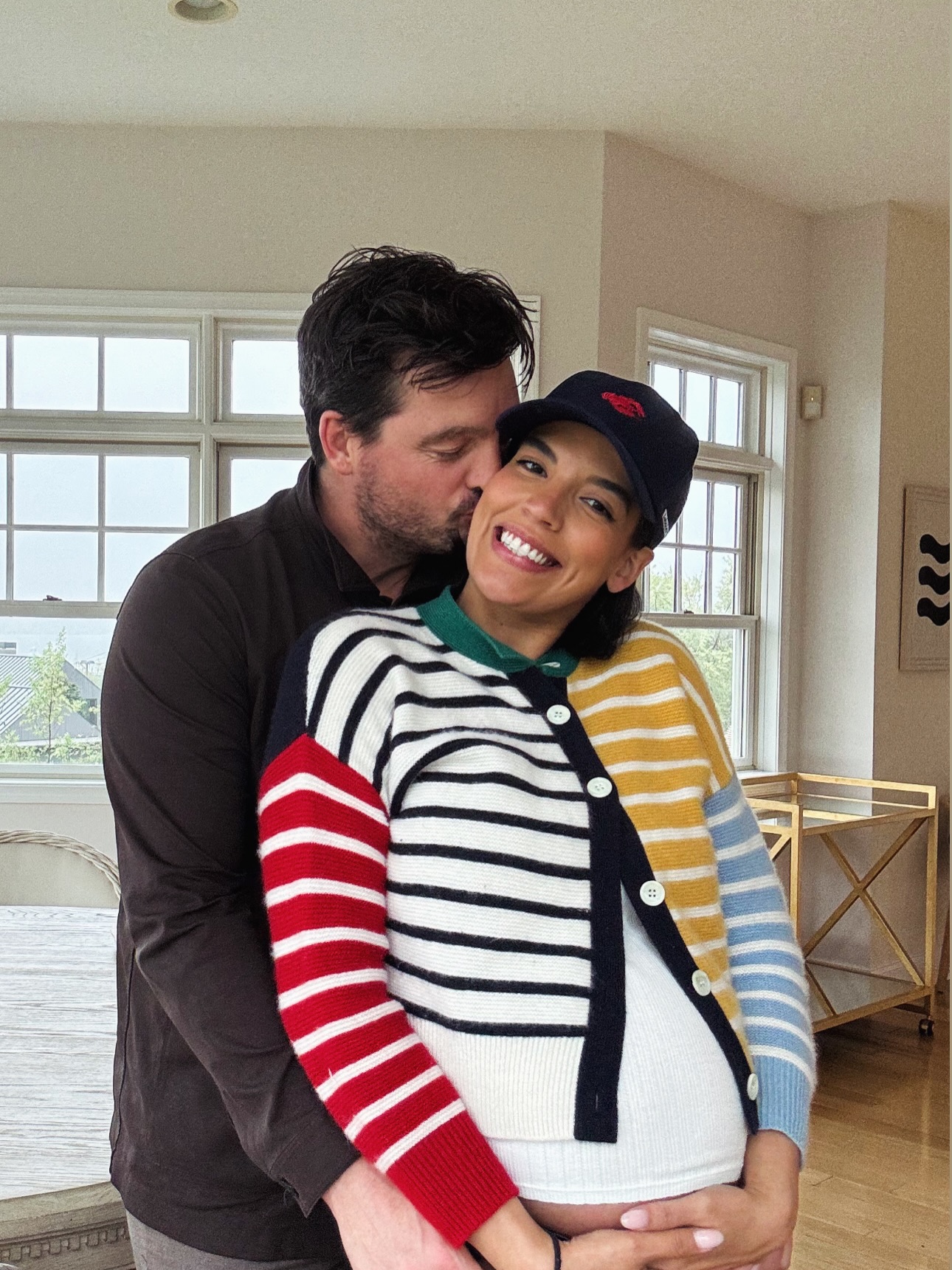 A man kisses Danielle Olivera's cheek, who smiles and wears a colorful striped sweater, while Eoin Heavey embraces her baby bump.