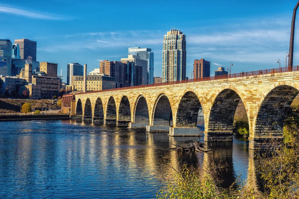 The Stone Arch Bridge, a historic stone arch bridge, spans the Mississippi River with the downtown Minneapolis skyline in the background.