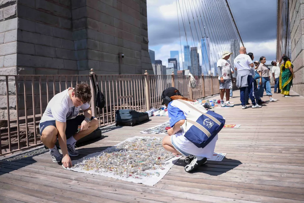 Two street vendors crouch over souvenirs displayed on the Brooklyn Bridge walkway, with New York City skyscrapers in the background.