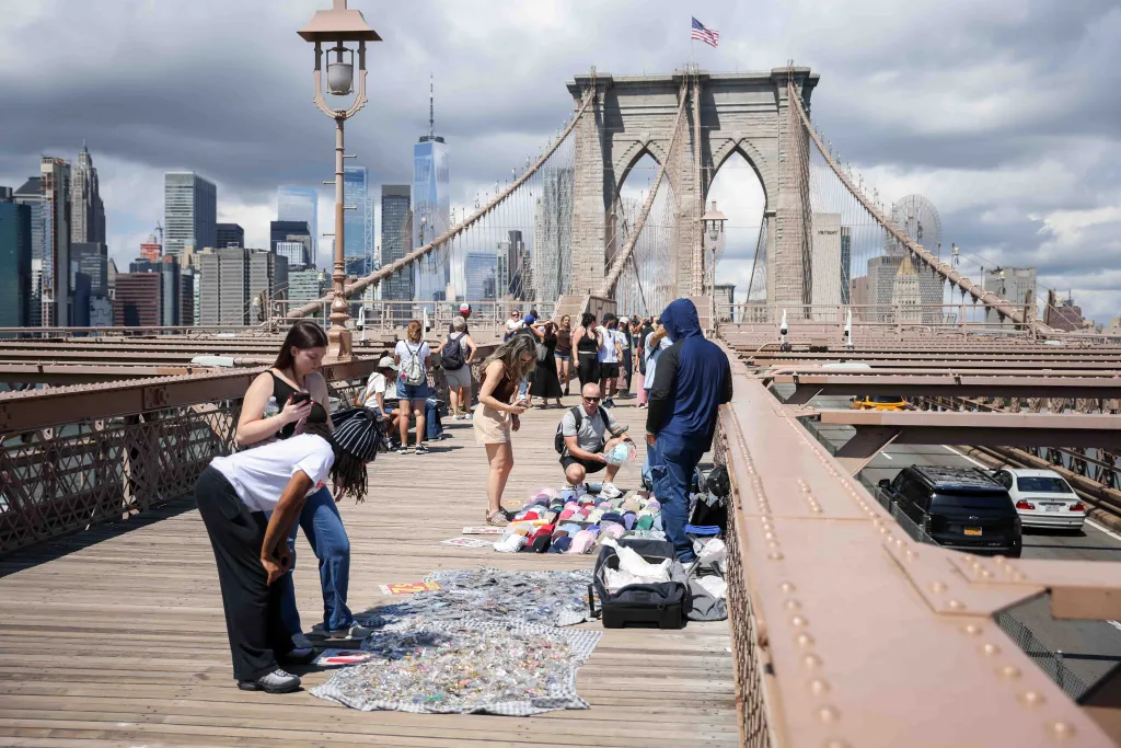 Street vendors selling souvenirs on the Brooklyn Bridge walkway with the New York City skyline in the background.
