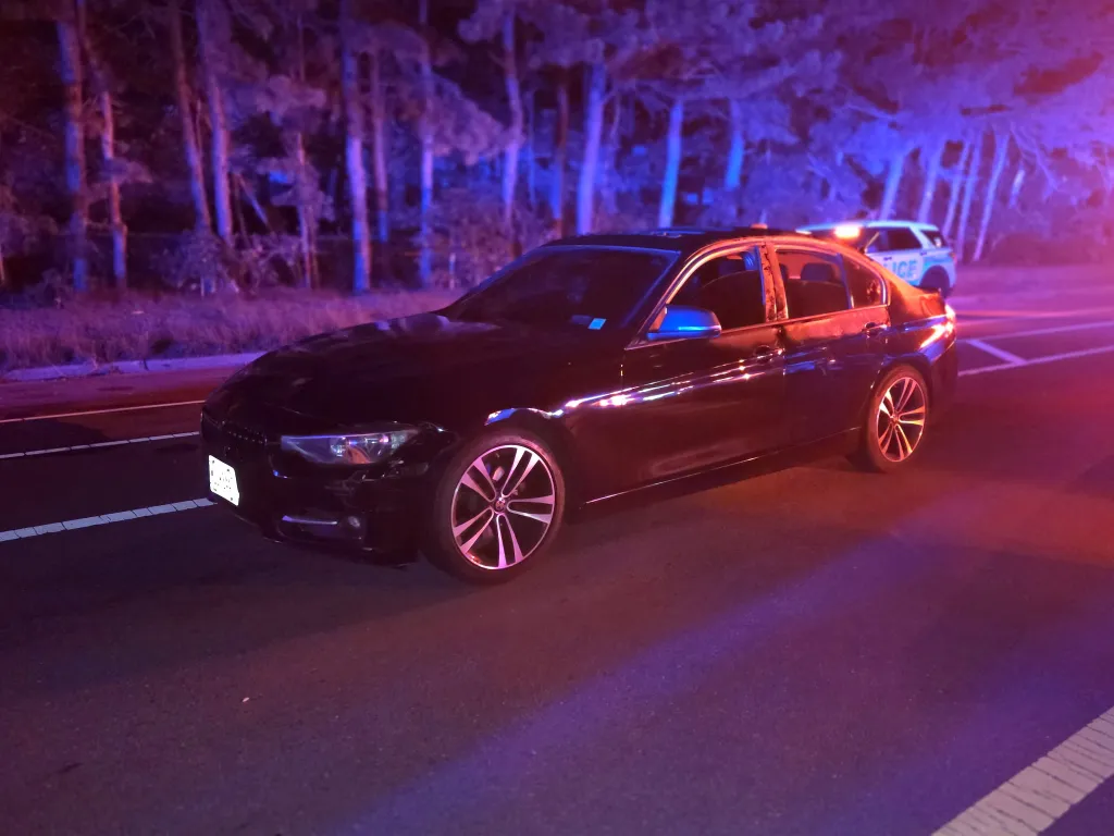 Black sedan on a road at night with red and blue police lights reflecting off its surface.