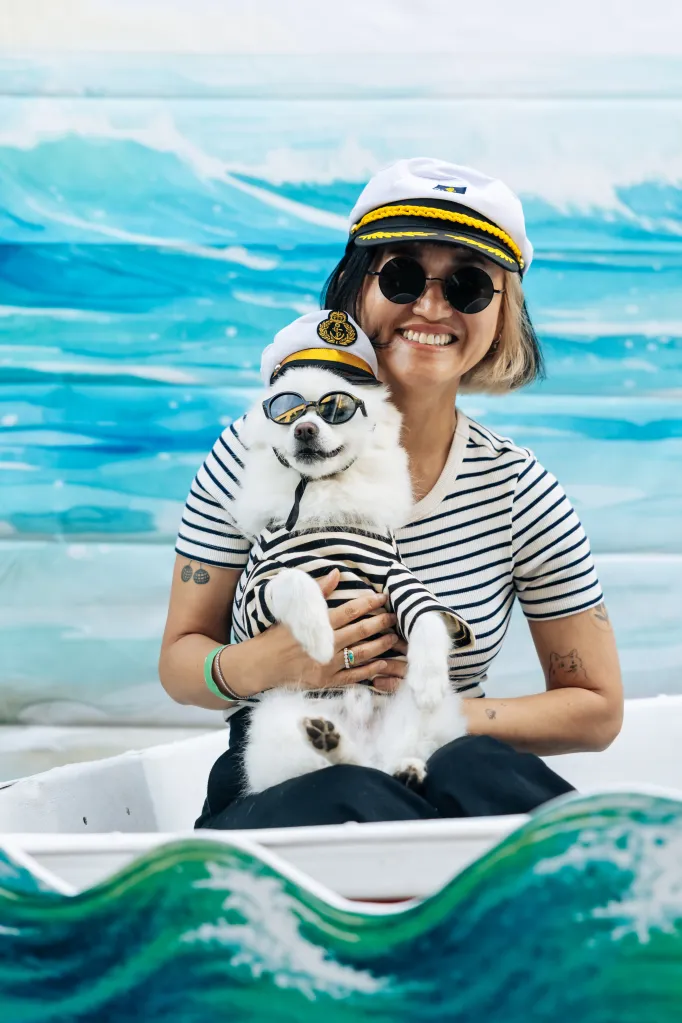 Suki Kwon, wearing a captain's hat and striped shirt, holds her dog Kumo, who is also wearing a captain's hat, sunglasses, and a striped shirt, in a boat with a painted wave backdrop.