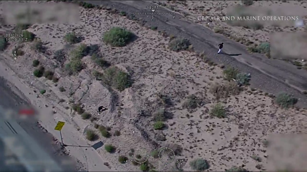 Aerial view of two individuals on foot, one running and one lying on the ground, in a desert landscape next to a road, tracked by 