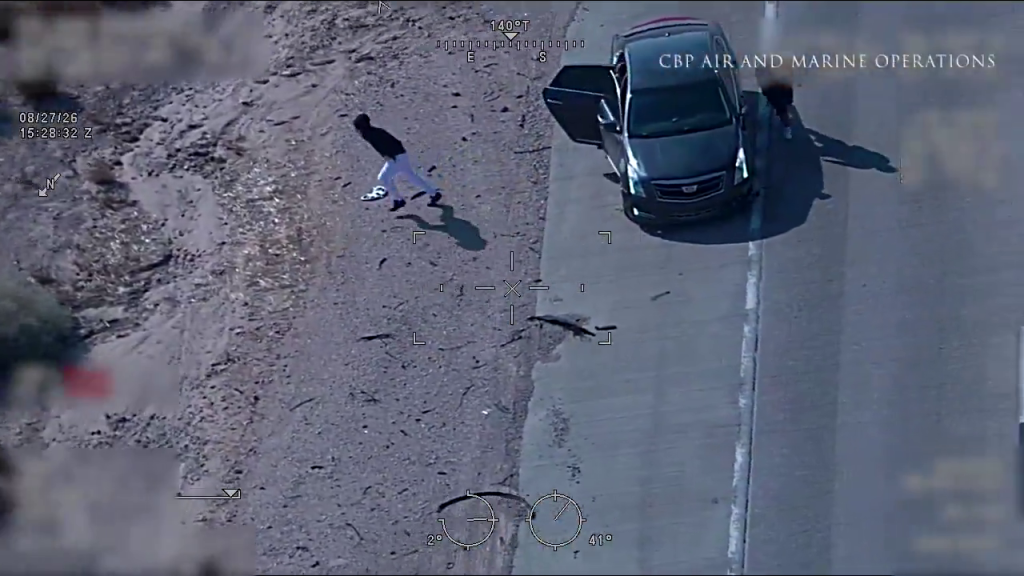 Aerial view of two people fleeing a dark sedan on a dirt shoulder next to a road, with 