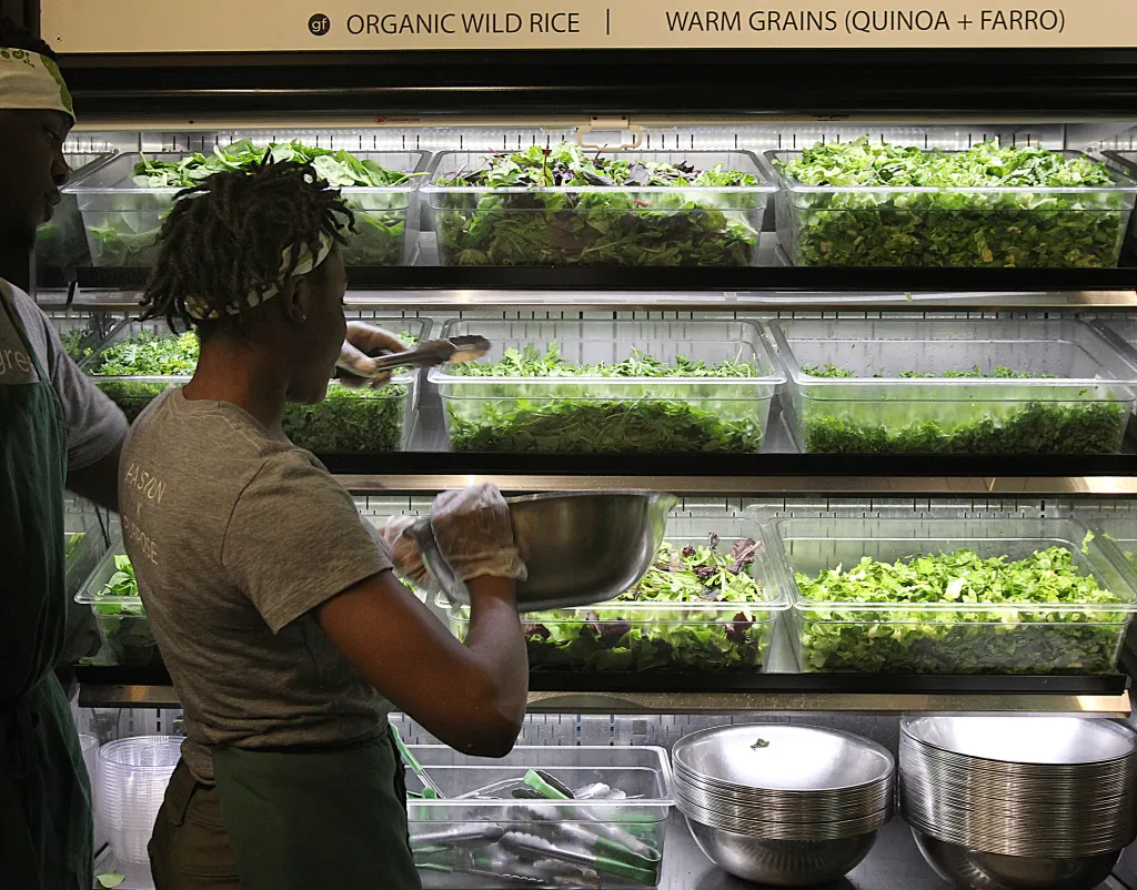 Employee at Sweetgreen salad bar prepares a salad from various greens in clear bins.