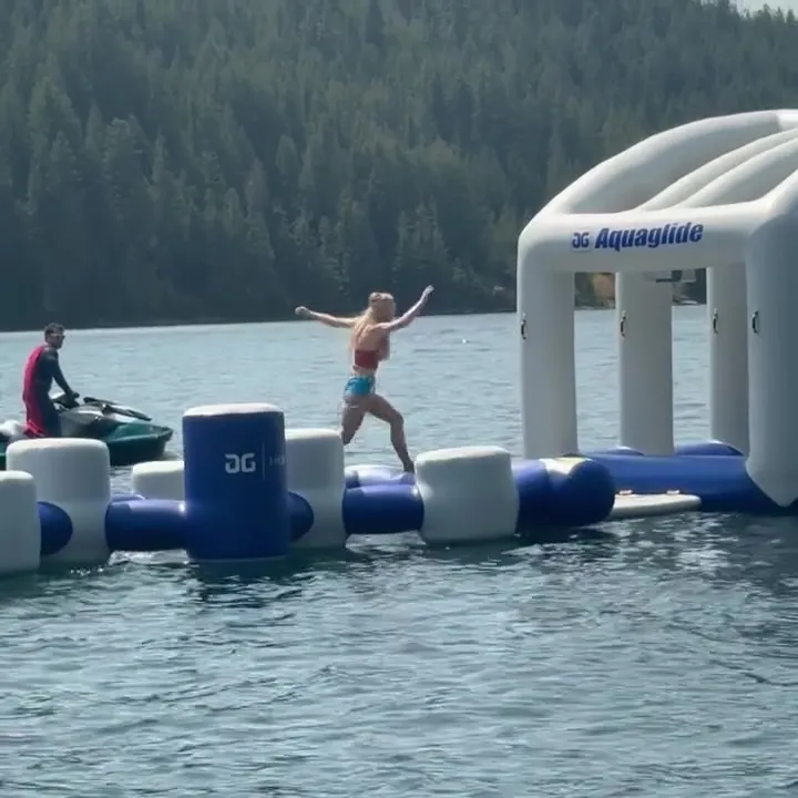 Sydney Sweeney in a Wonder Woman costume playing on an inflatable water obstacle course with Scooter Braun in a Superman costume on a jet ski behind her.