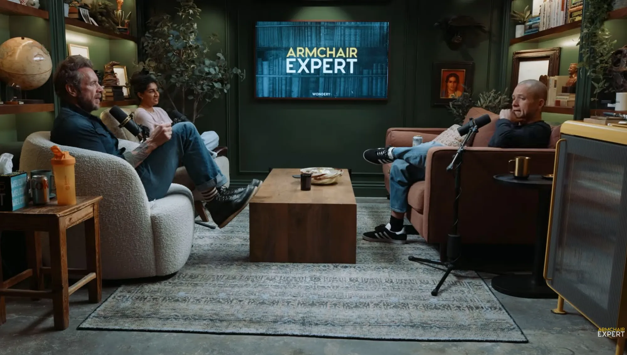 Three people sitting in armchairs, two men and one woman, conversing in a podcast studio with a screen displaying 