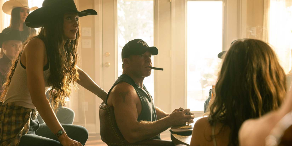 Taylor Sheridan as Travis Wheatley in Yellowstone, smoking a cigar, with a woman beside him. He wears a black baseball cap and black tank top.