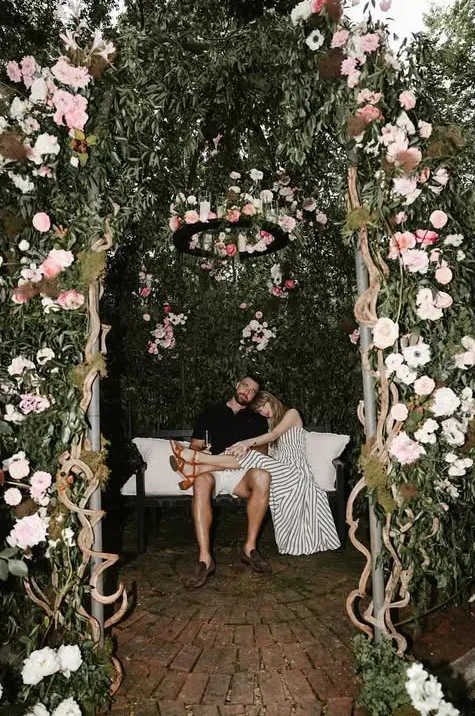 Taylor Swift sitting with Travis Kelce on a bench under a floral archway.