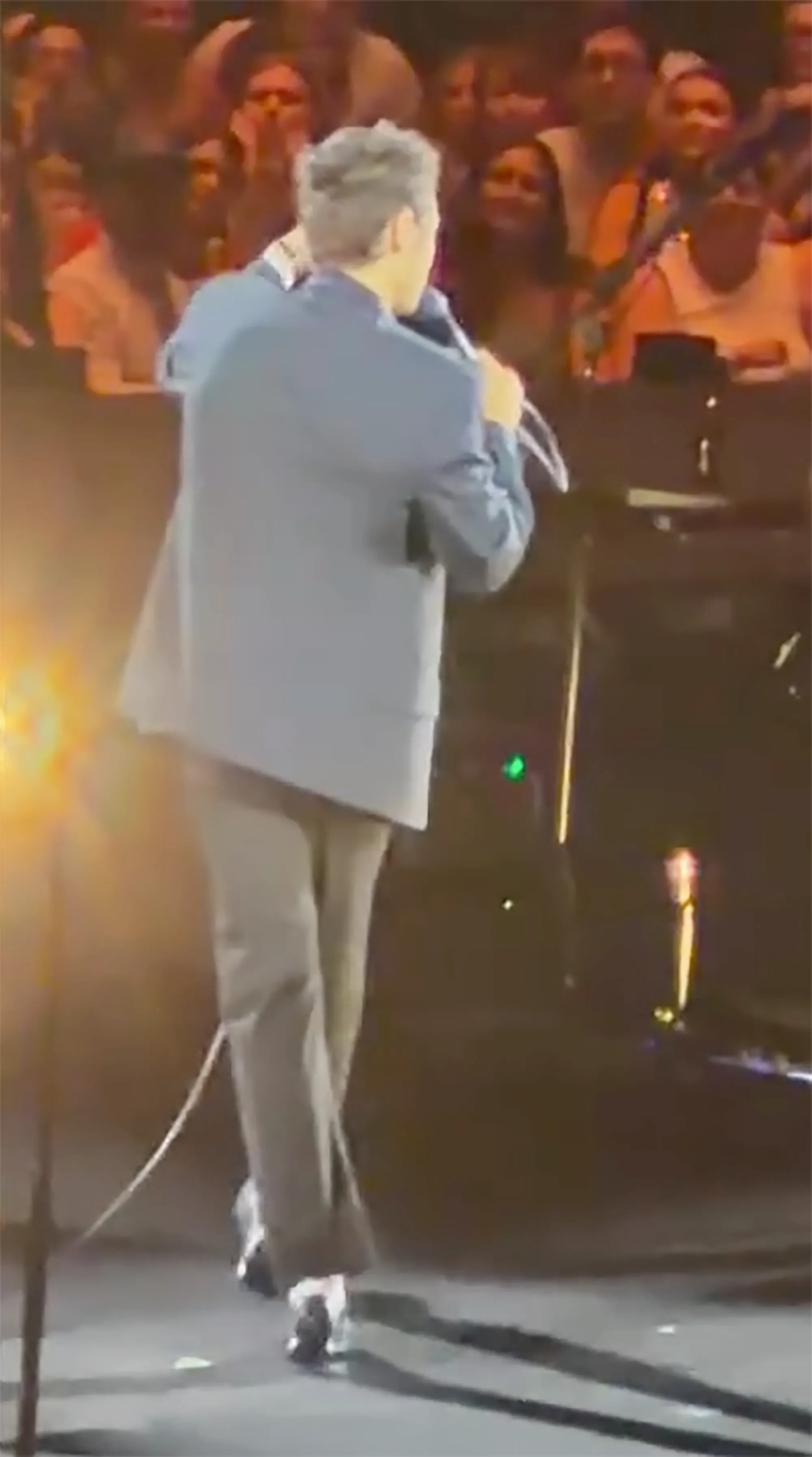 A man in a suit jacket with his back to the viewer stands on a stage with a microphone in hand, facing a crowd.