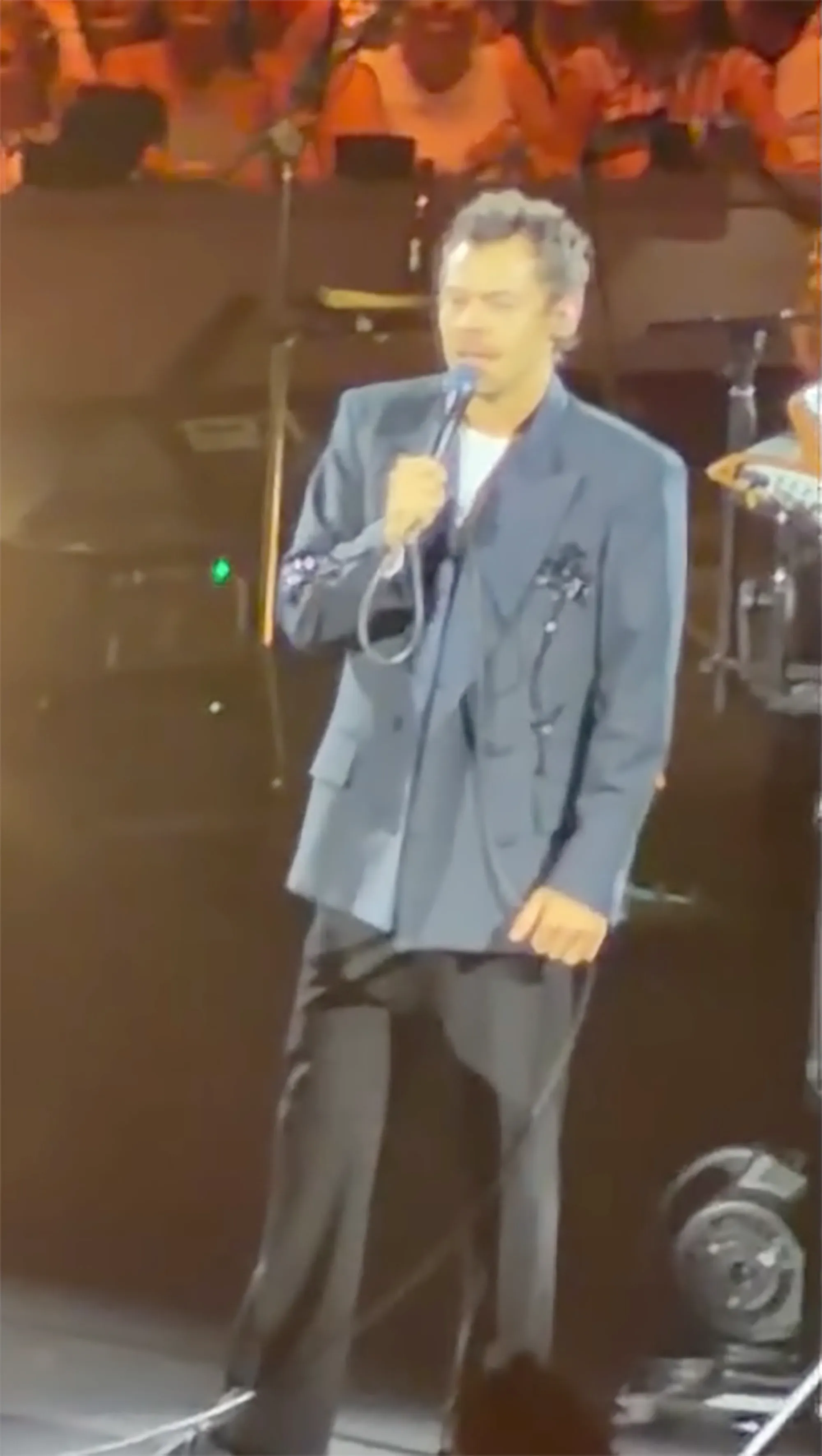 Harry Styles on stage, wearing a gray suit jacket over a white t-shirt, holding a microphone.