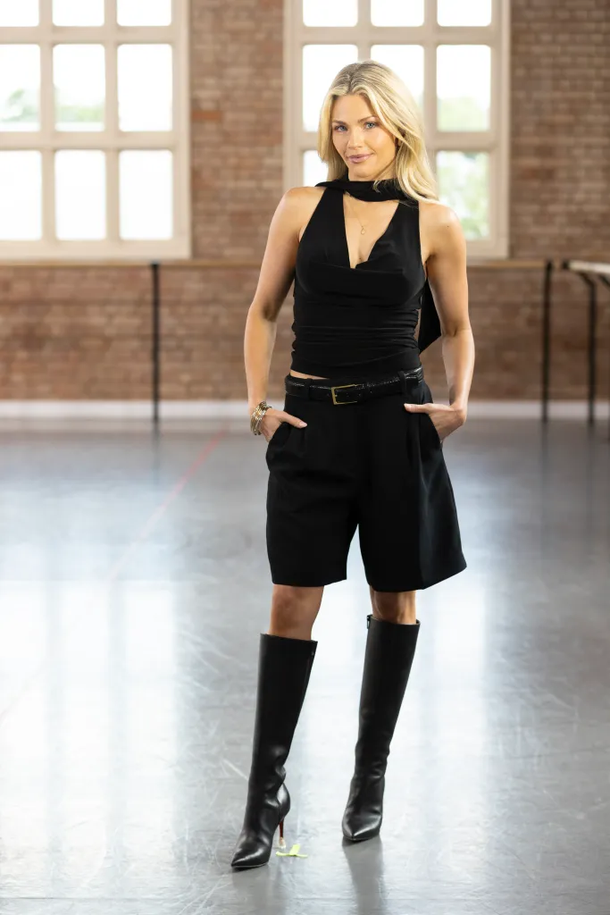 Witney Carson poses in black attire, including a draped top, shorts, and knee-high boots, in a dance studio.