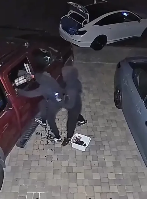 Security camera footage of two hooded figures removing items from a vehicle.