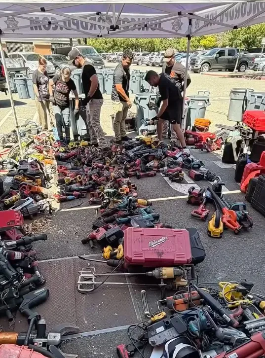 Police officers inspect a large collection of recovered power tools spread across a parking lot.