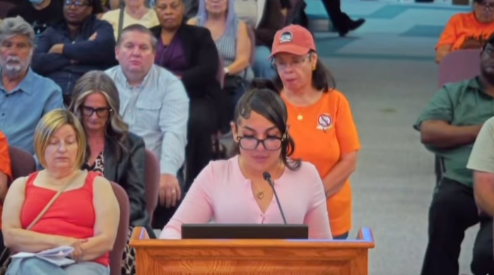Teen activist Sadie, wearing glasses and a light pink shirt, speaks at a podium with a microphone.