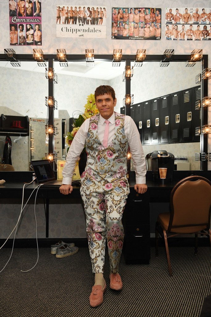 Perez Hilton in a floral suit with a pink tie, leaning on a vanity table with Chippendales posters on the wall.