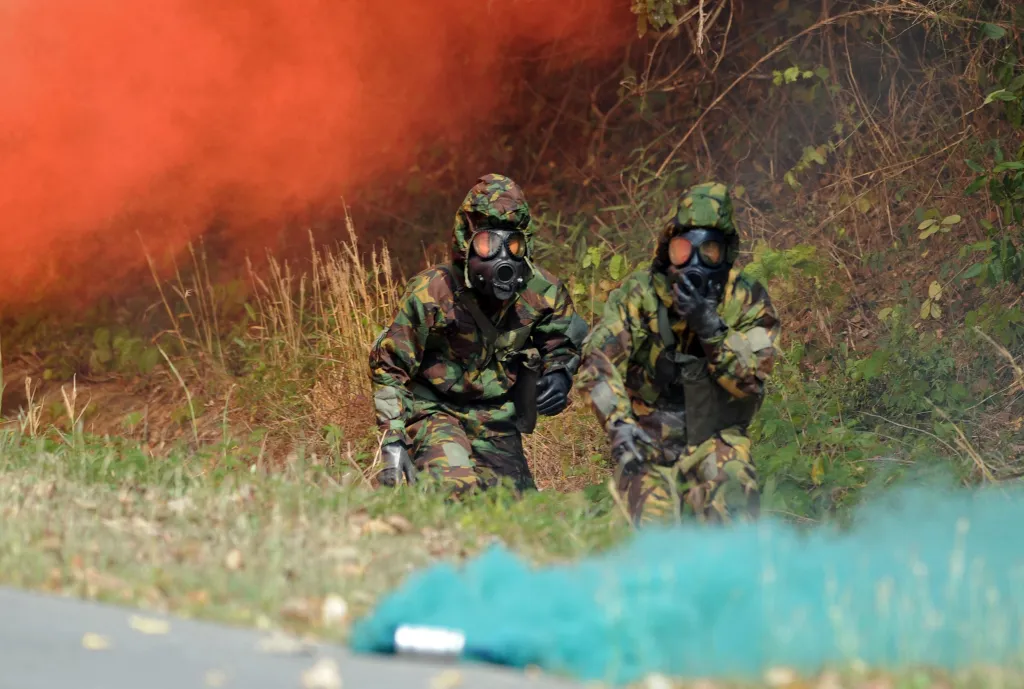 Two soldiers in camouflage and gas masks walk through red and blue smoke during a military exercise.