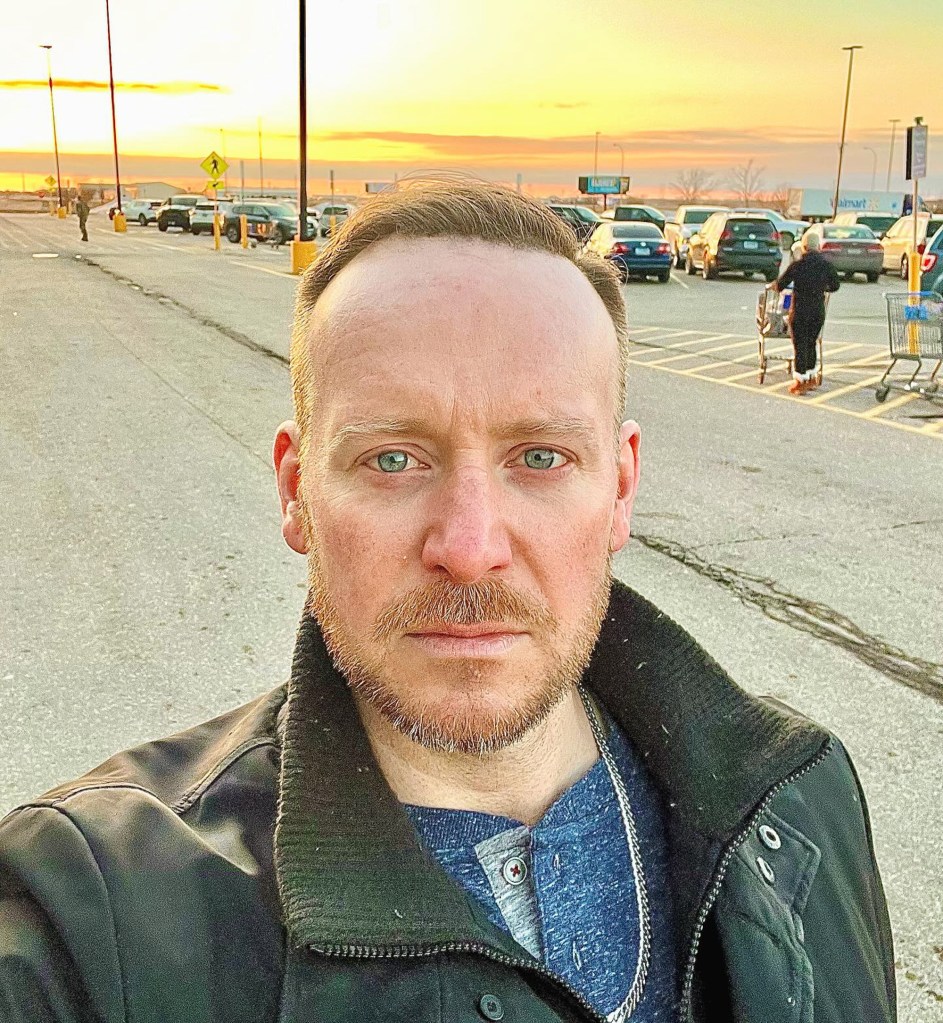 Tyler Duckworth in a parking lot with cars and a bright sunset in the background.