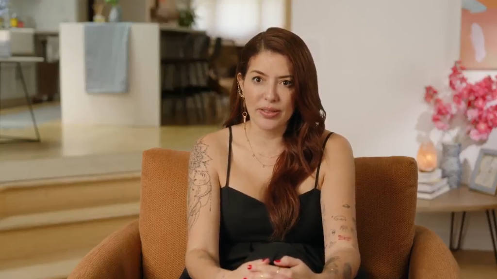 Paula from 90 Day Fiance, with long reddish-brown hair and tattoos on her arms, sits in an armchair wearing a black dress.
