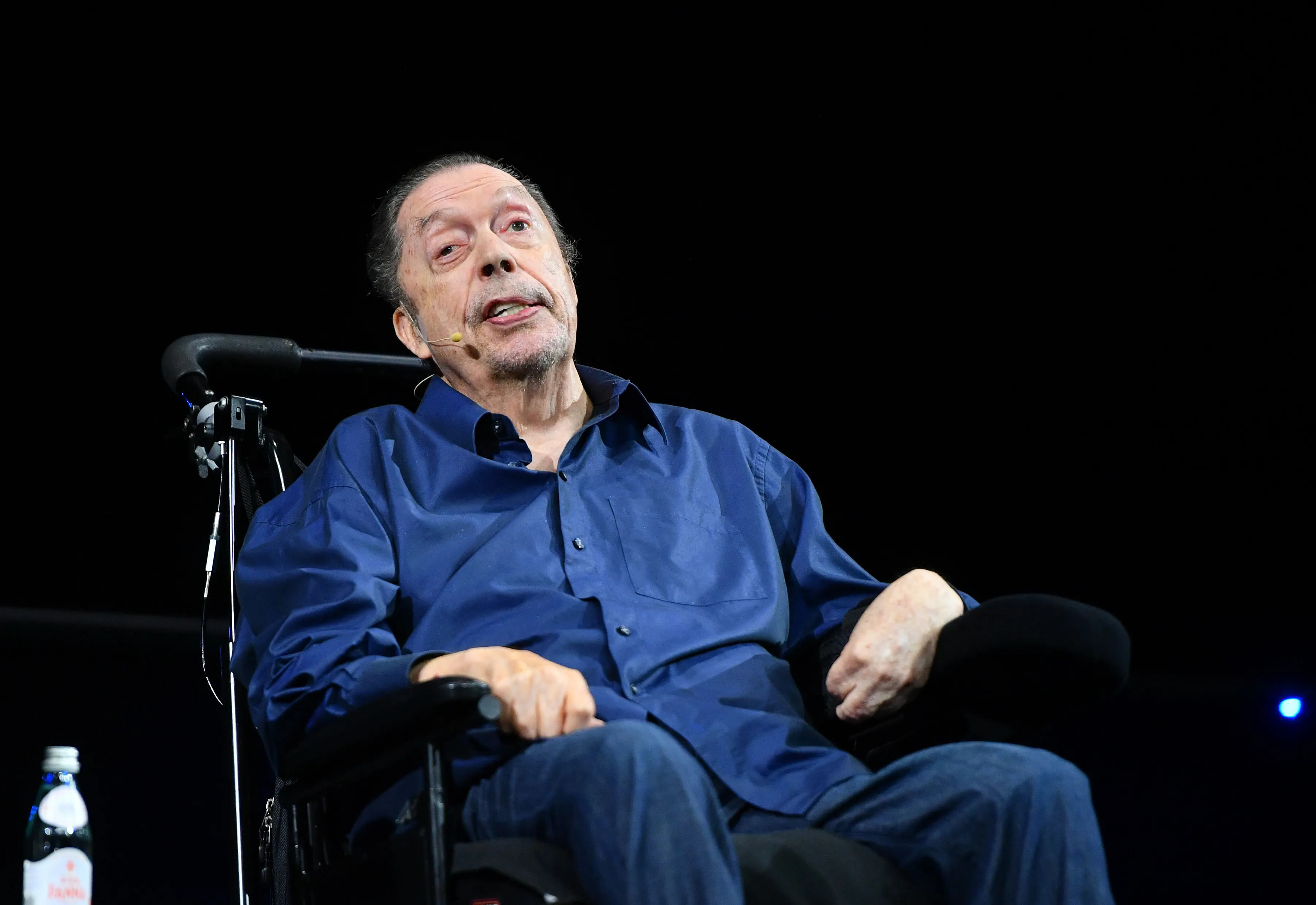 Tim Curry in a wheelchair, wearing a blue shirt and jeans, speaks with a microphone near his mouth at the 50th Anniversary of