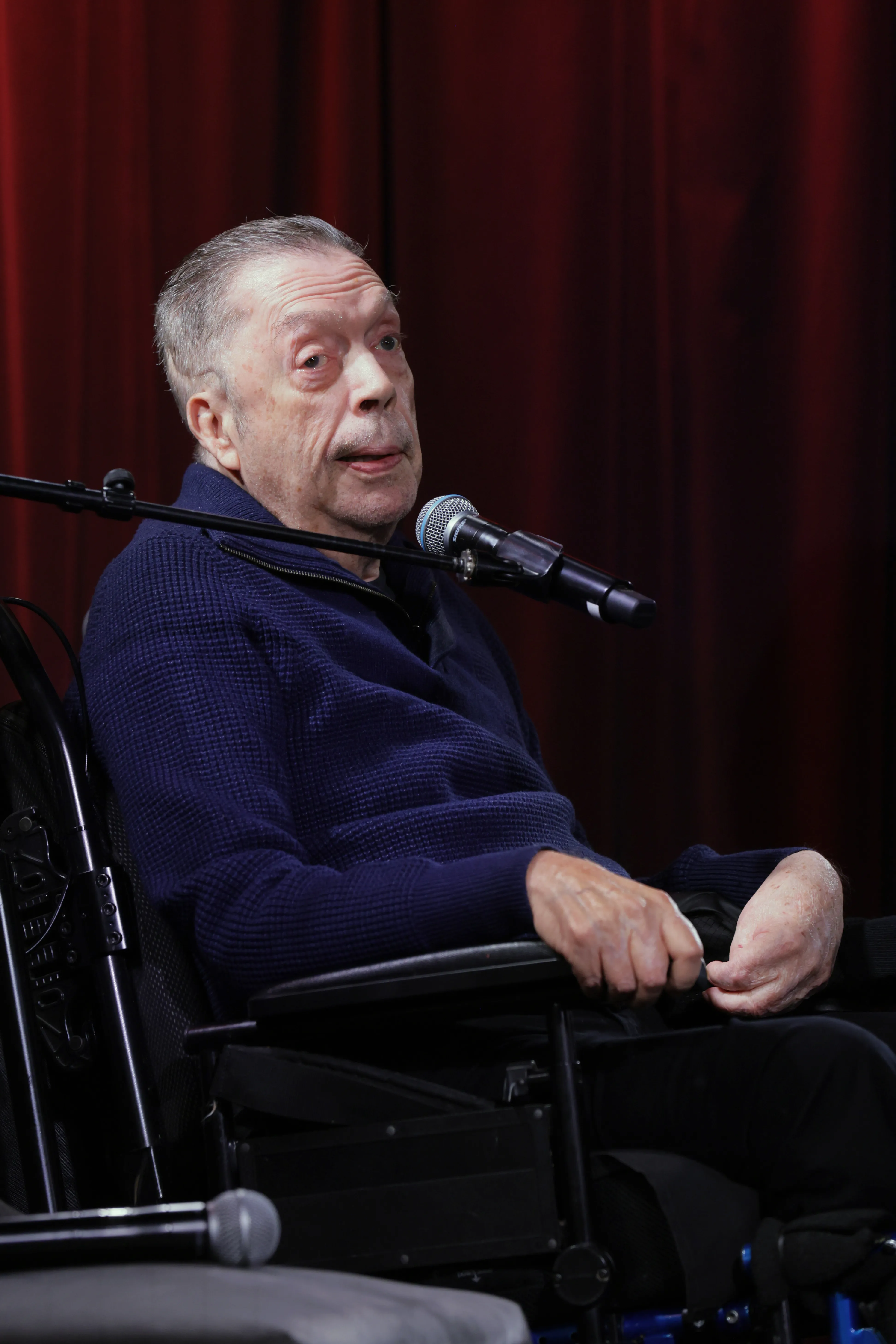 Tim Curry in a wheelchair speaking onstage at the GRAMMY Museum L.A. Live.