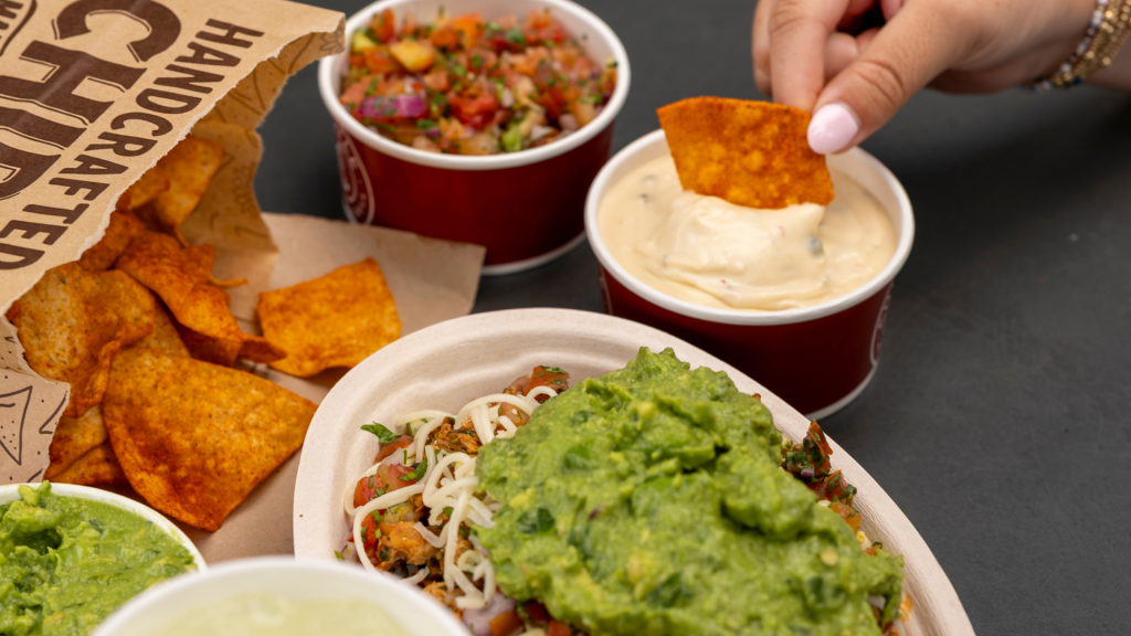 A hand dipping a Chili Lime Chip into a cup of queso, alongside a Pollo Asado bowl and other Chipotle items.