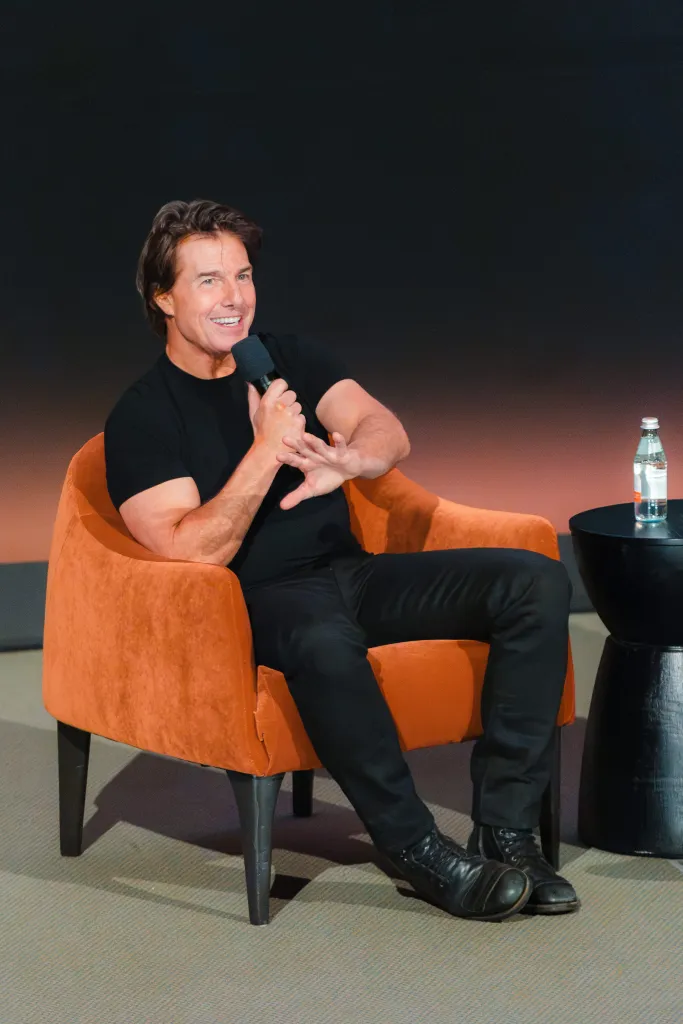 Tom Cruise speaking into a microphone while seated on an orange chair.