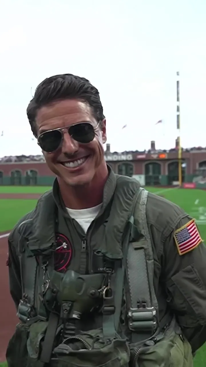 Jerome LeBlanc, a Tom Cruise look-alike dressed as Maverick from Top Gun, smiles in an olive green flight suit and aviator sunglasses with a baseball field in the background.