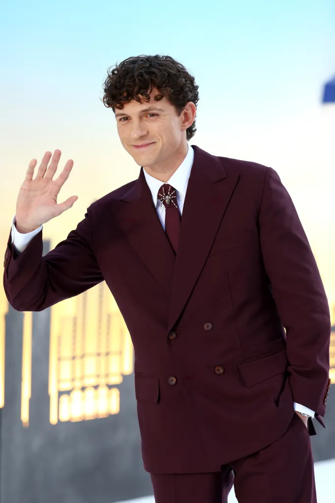 Tom Holland, in a maroon suit with a spider pin on his tie, smiles and waves.