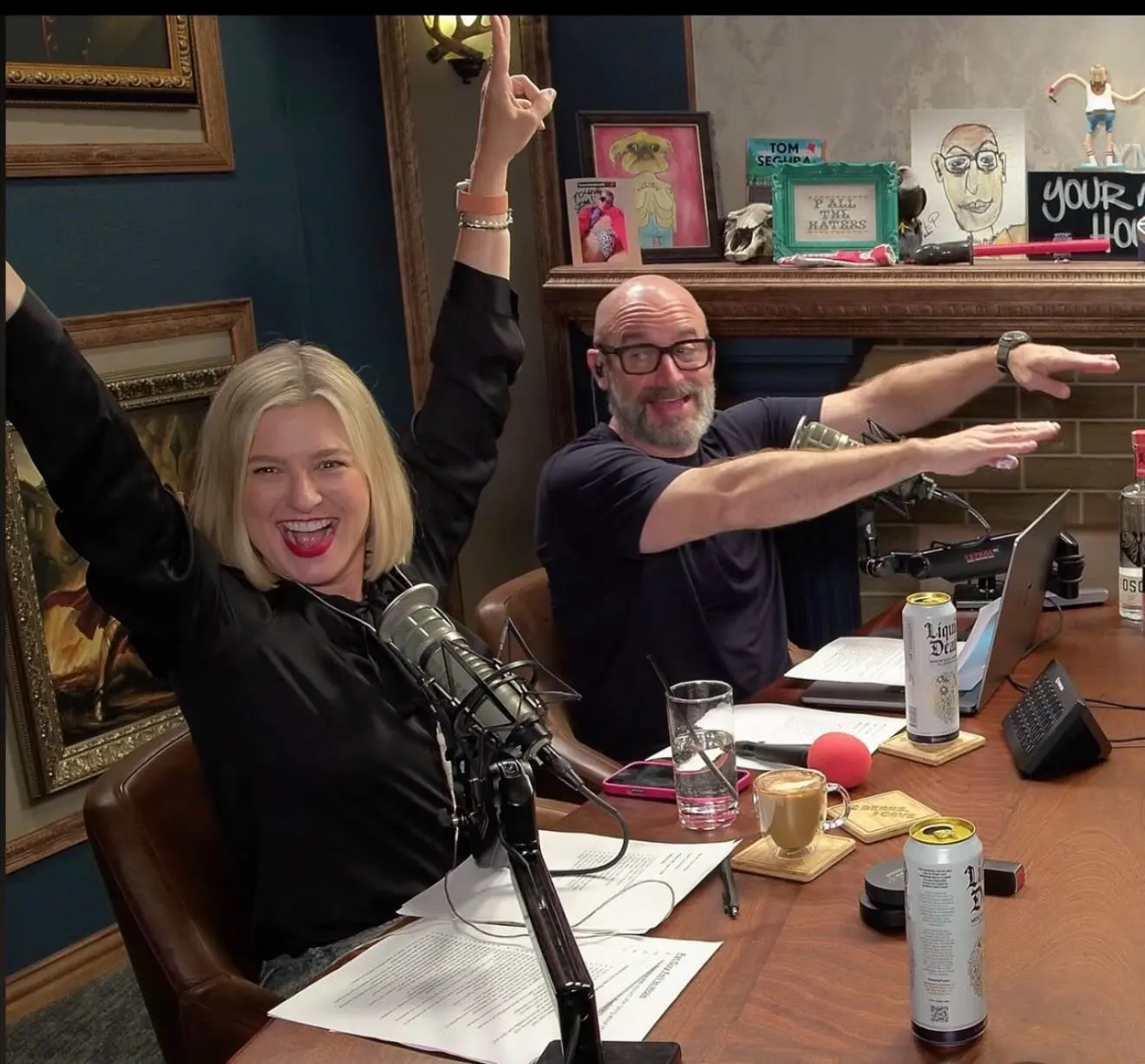 Tom Segura and Christina Pazsitzky raising their arms with excitement while seated at a podcasting desk.