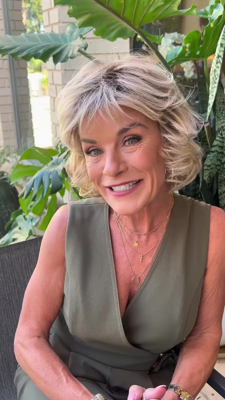Toni Brattin, wig designer, smiling and wearing a green sleeveless top and multiple gold chain necklaces.