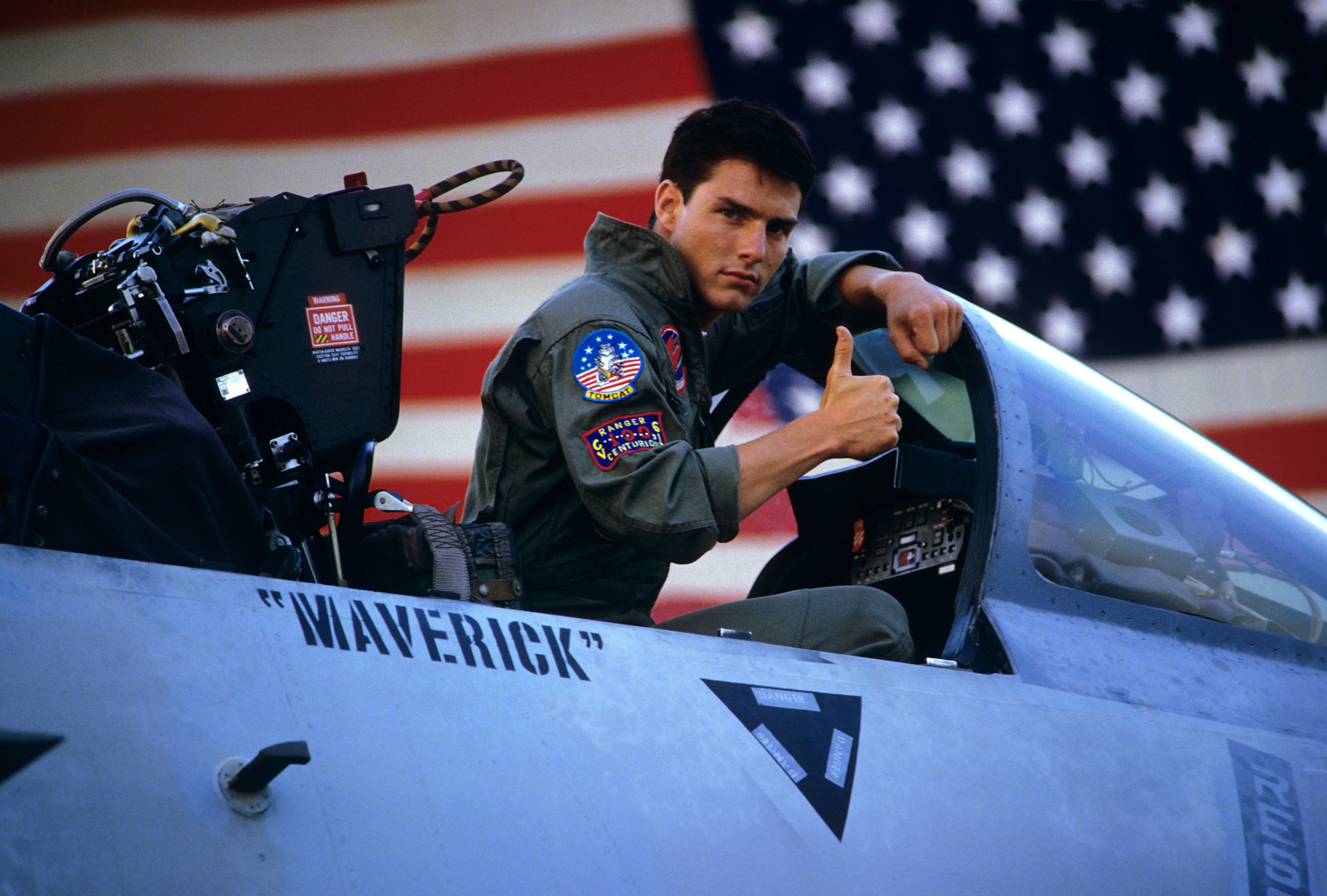 Tom Cruise as Maverick giving a thumbs up in the cockpit of a fighter jet with an American flag in the background.