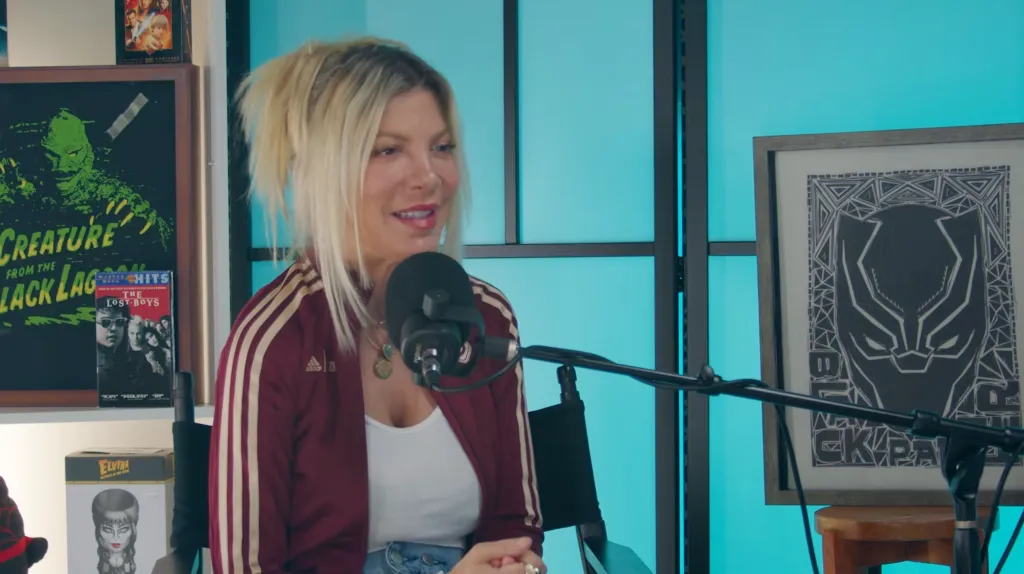 Tori Spelling speaking into a microphone while wearing a maroon track jacket with white stripes.
