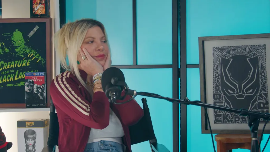 Tori Spelling resting her face in her hands while seated in front of a microphone.